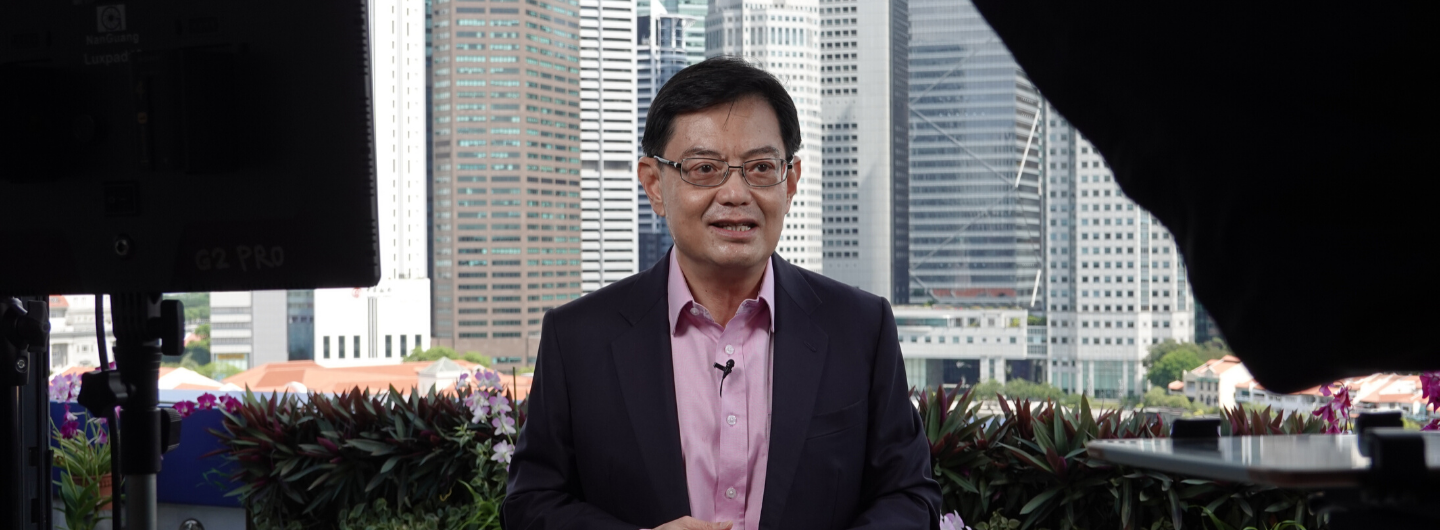 Heng Swee Keat, in glasses and suit, speaks in front of a skyline and filming equipment.